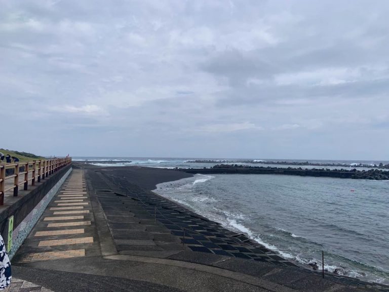 曇り空の下、黒い砂浜と護岸のむこうで白波が砕ける八丈島の海岸