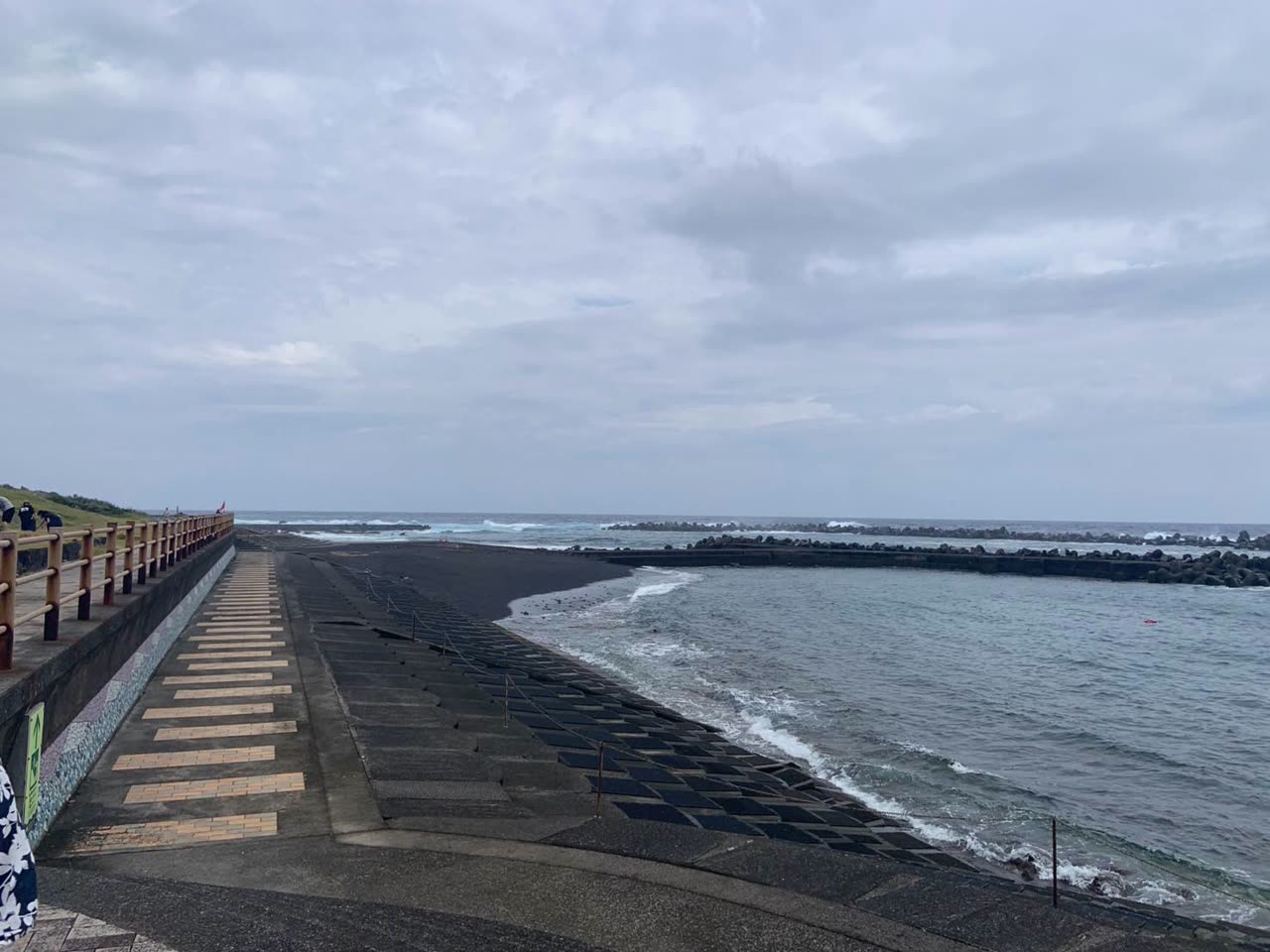 曇り空の下、黒い砂浜と護岸のむこうで白波が砕ける八丈島の海岸