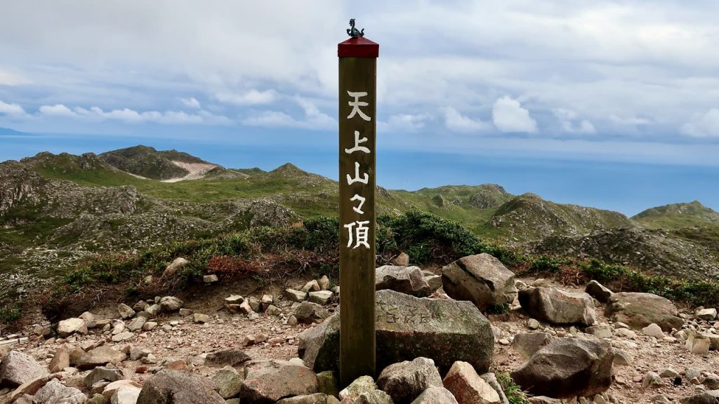 「天上山々頂」と刻まれた木の標柱。岩がごろごろした山頂部と、遠くに海が見える
