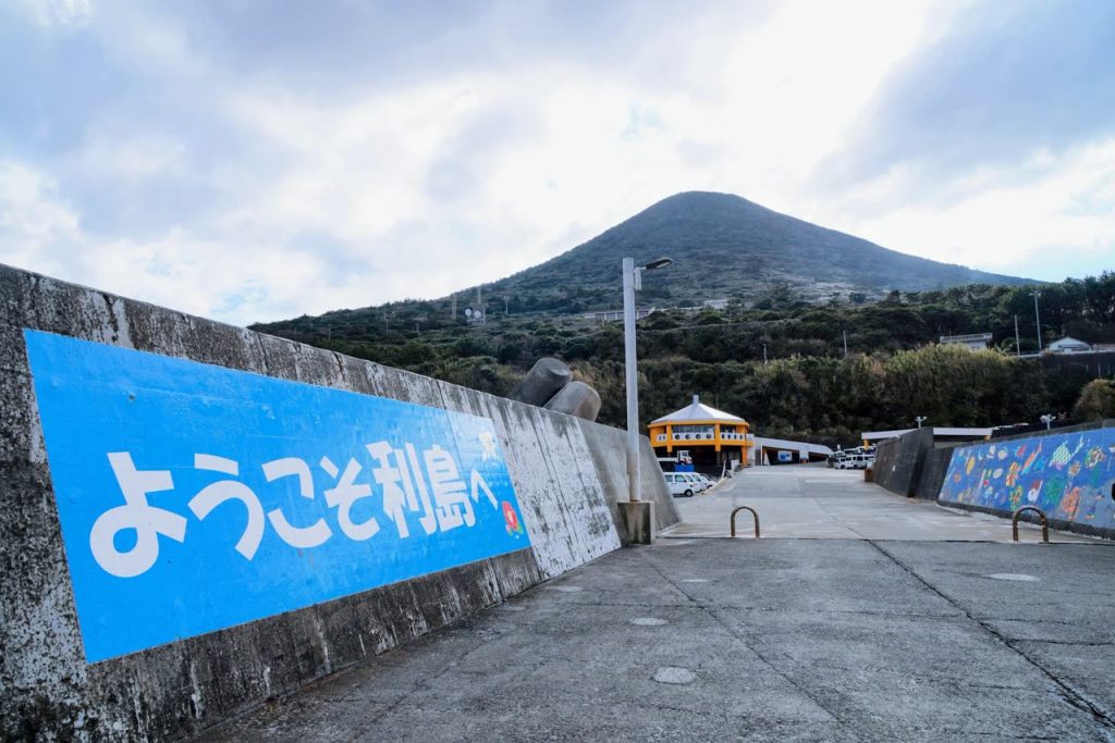 港の「ようこそ利島へ」の看板と、円錐形の宮塚山